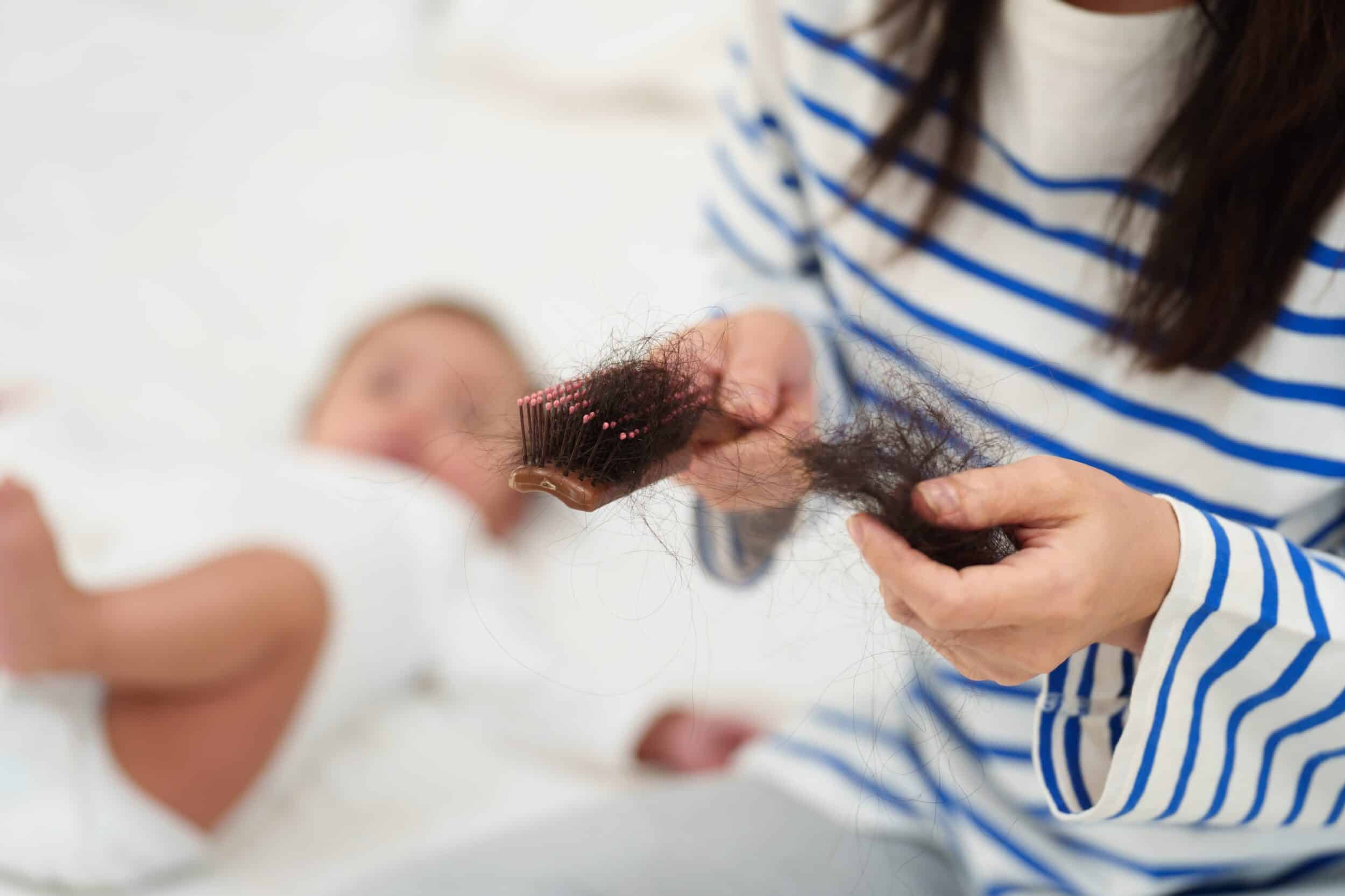 Woman sitting next to her newborn baby finds hair in her brush, a sign of postpartum hair loss.