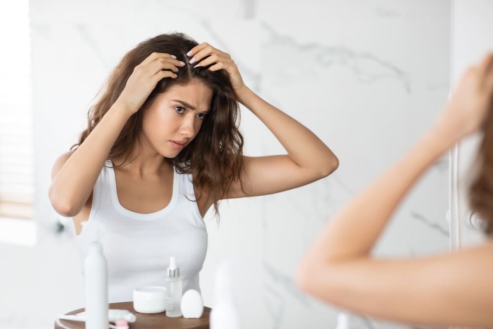 Woman looking in the mirror touching her head as she notices hair loss.